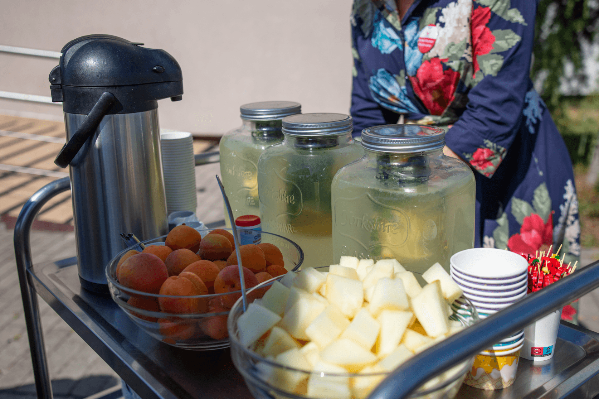 Căruț de servire cu trei borcane mari de limonadă cu mentă, un bol cu caise proaspete și un bol cu bucăți de pepene galben, alături de pahare și un termos, servite în aer liber de o îngrijitoare în rochie florală