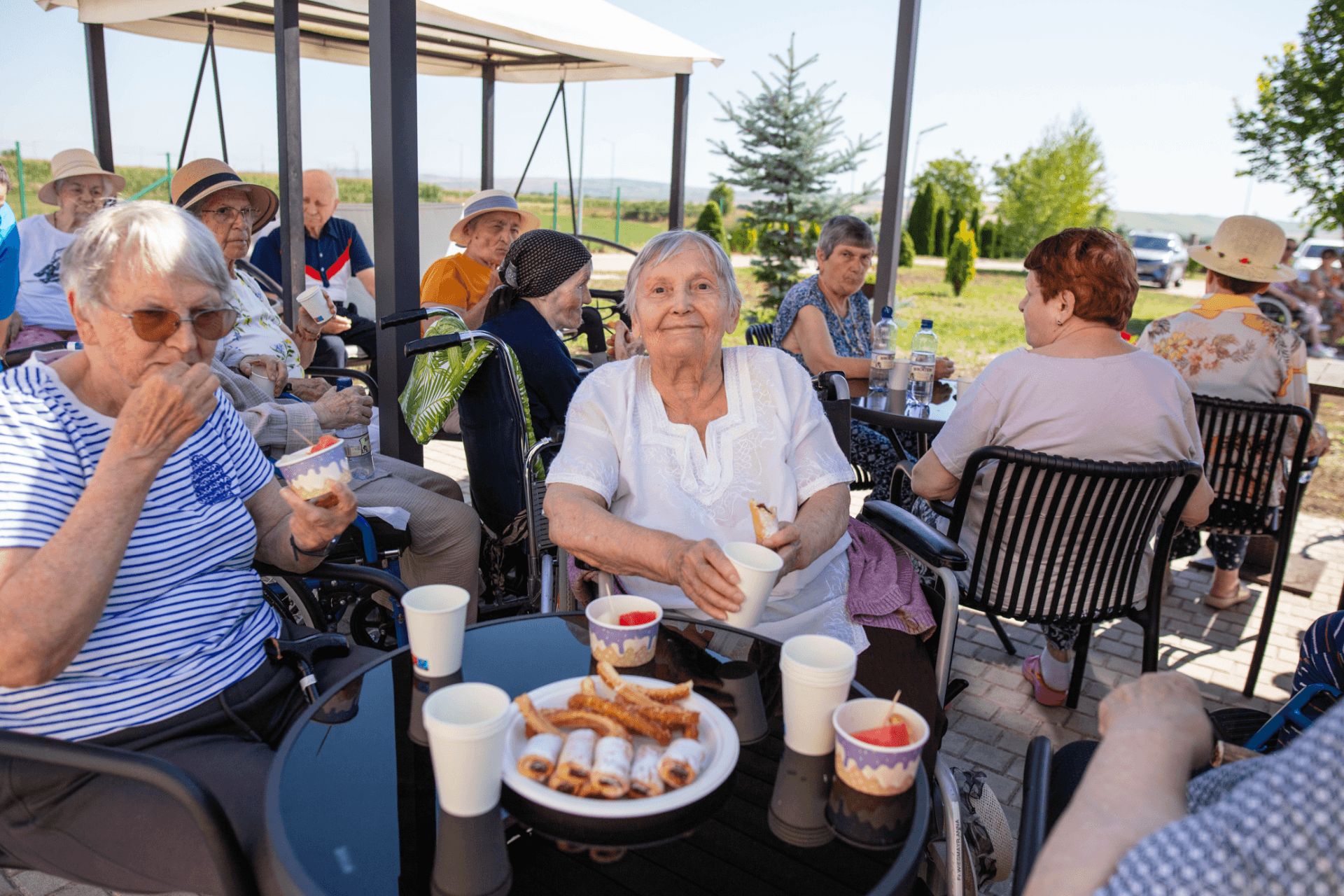 Grup de bătrâne așezate la mese în aer liber, bucurându-se de gustări și băuturi într-un centru de îngrijire seniori din România, pe vreme însorită