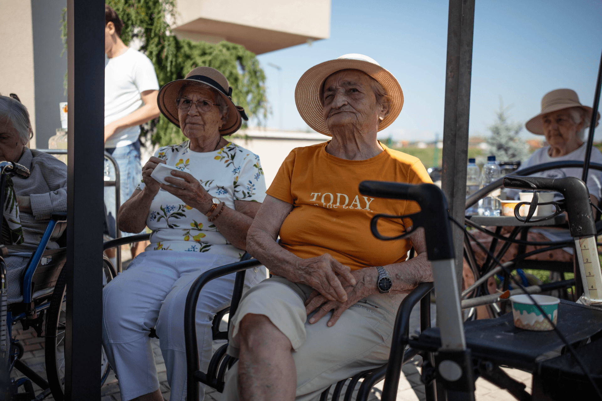 Două doamne în vârstă cu pălării de soare stau pe scaune afară la un centru de îngrijire pentru seniori, una purtând un tricou portocaliu cu inscripția TODAY