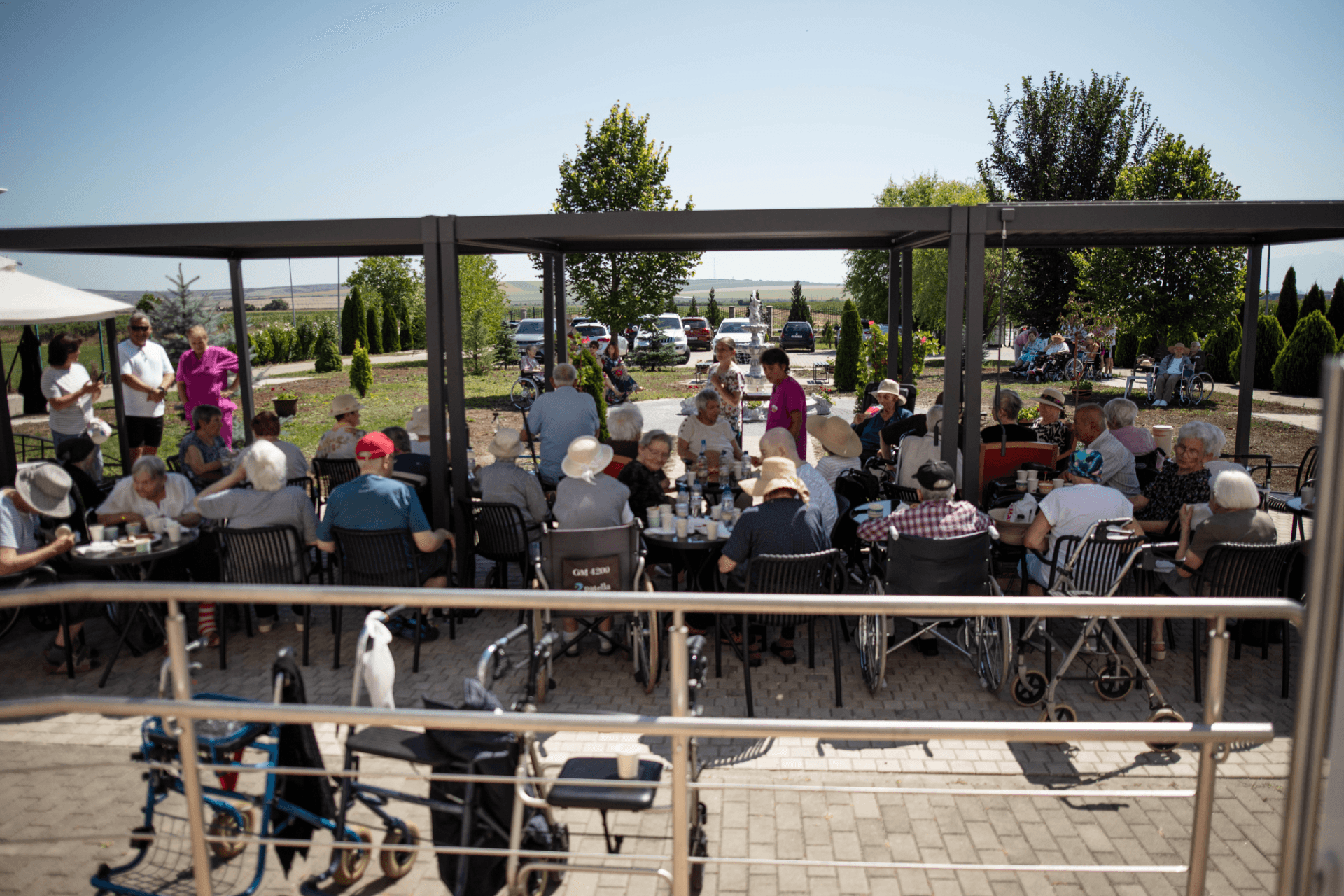 Grup de seniori așezați la mese sub un foișor exterior, participând la o activitate socială în cadrul unui centru de îngrijire din România, unii folosind scaune cu rotile