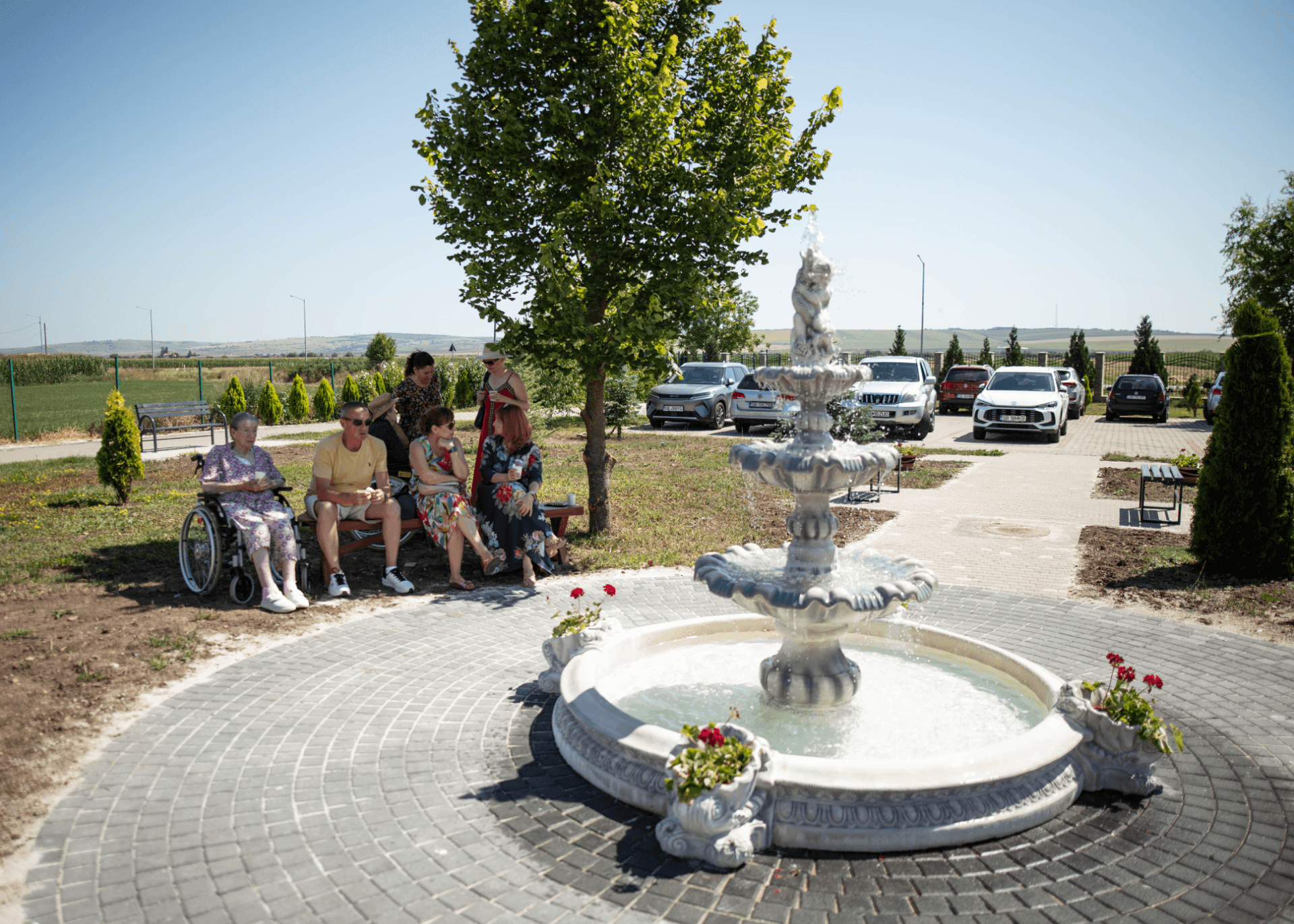 Bătrână în scaun rulant alături de familie pe o bancă în grădina centrului de îngrijire seniori, cu fântână arteziană albă în prim-plan și parcare în fundal