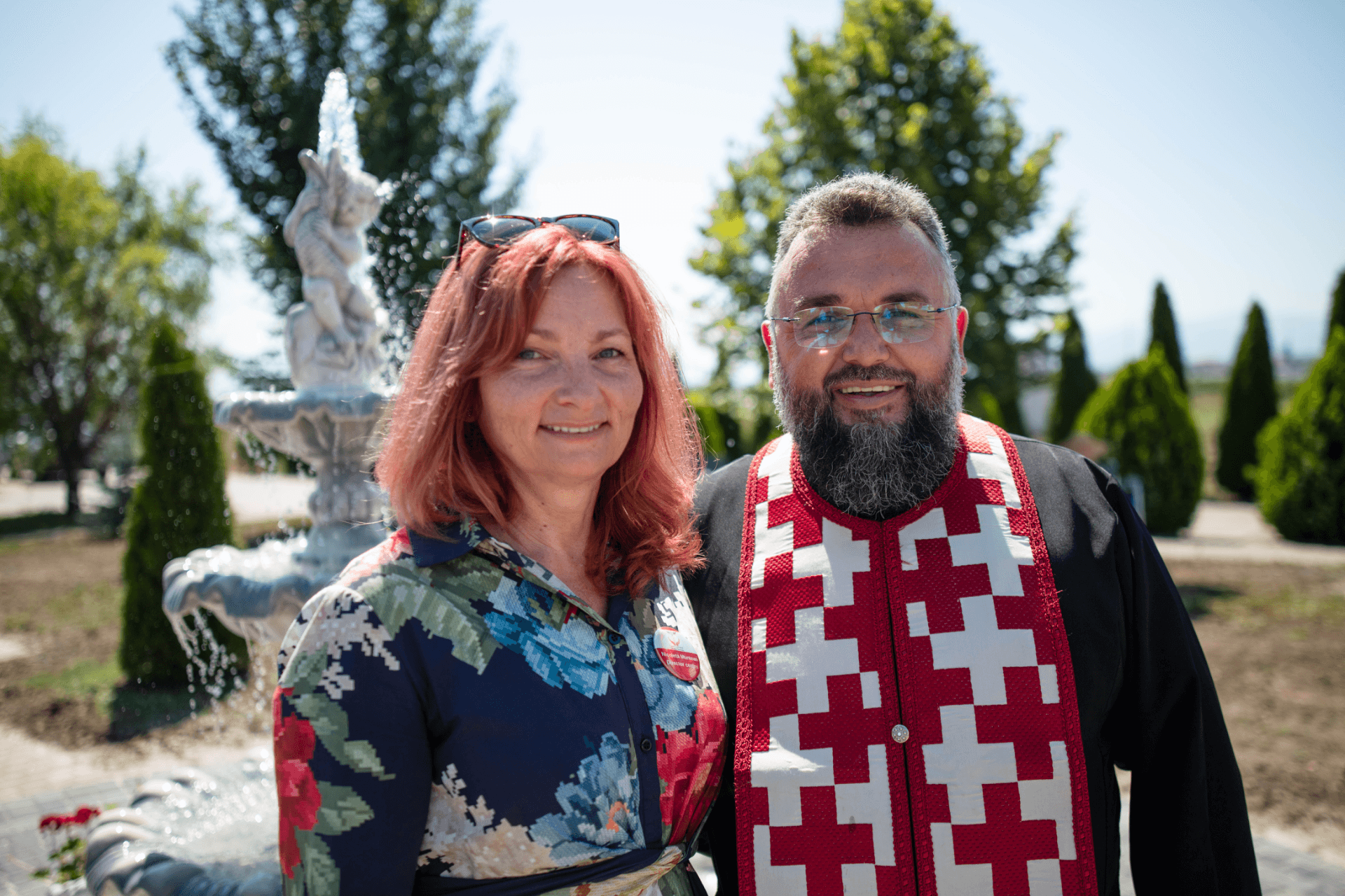 O femeie cu păr roșcat și un bărbat cu barbă purtând vestă tradițională roșie cu alb, stând împreună lângă o fântână artezică cu statui în grădina centrului de seniori