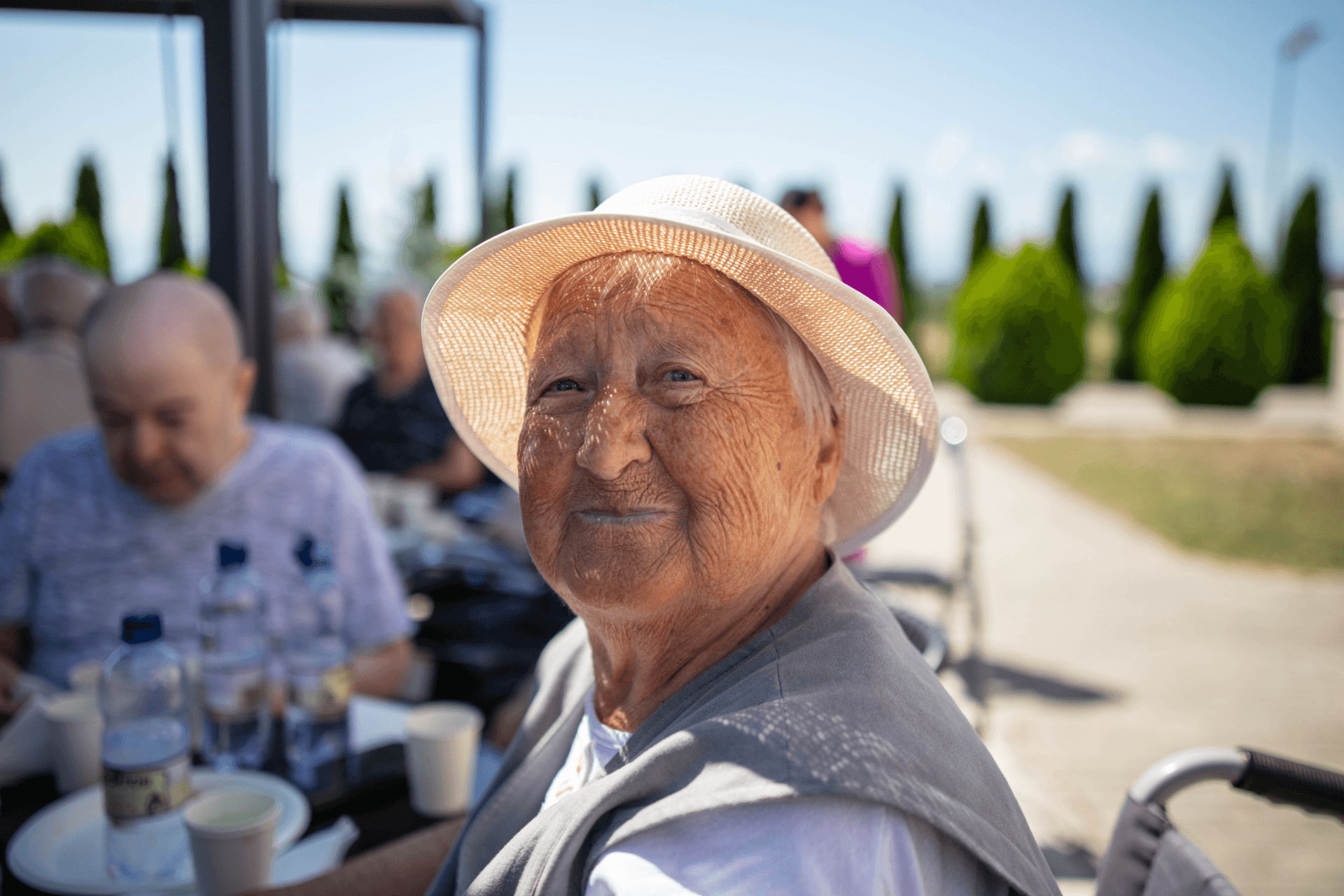 Femeie în vârstă cu pălărie albă de paie, zâmbind la cameră, așezată afară la masă în grădina unui centru de îngrijire pentru seniori, cu chiparoși în fundal