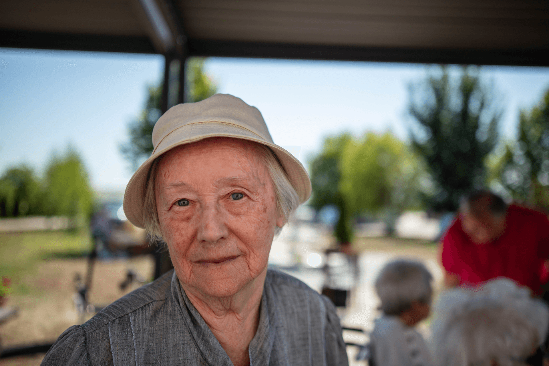 Femeie în vârstă purtând pălărie bej și bluză gri, fotografiată în exterior la un centru de îngrijire pentru seniori din România, cu alți rezidenți în fundal