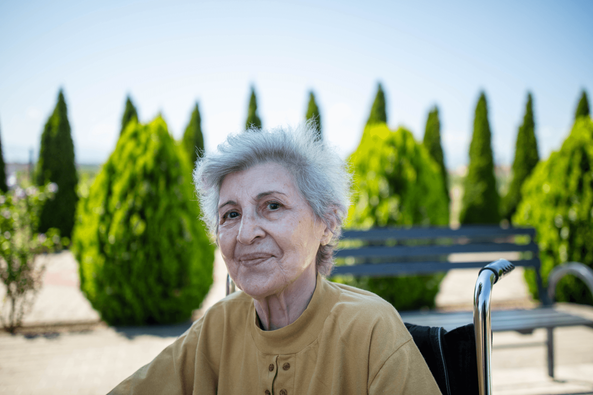 Femeie în vârstă cu păr alb și bluză bej, așezată în scaun cu rotile, zâmbind ușor în grădina centrului de îngrijire, cu brazi ornamentali în fundal